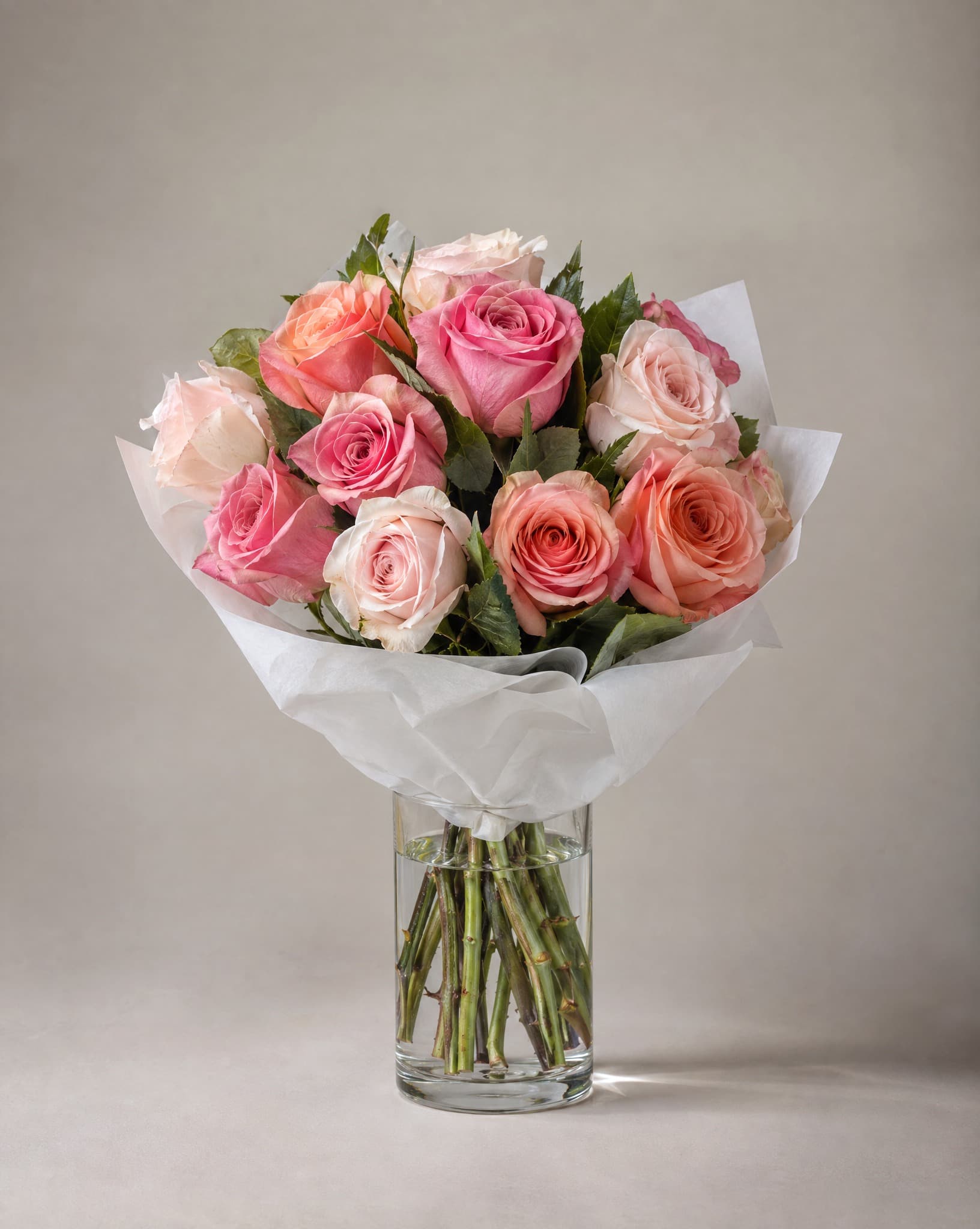 A dozen pink, coral and cream roses wrapped in white paper in a clear glass vase