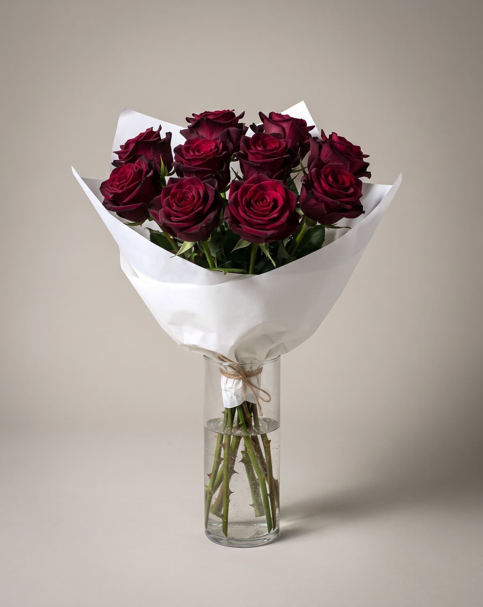 A dozen deep red roses hand-wrapped in white paper and tied with twine, standing in a tall clear glass vase