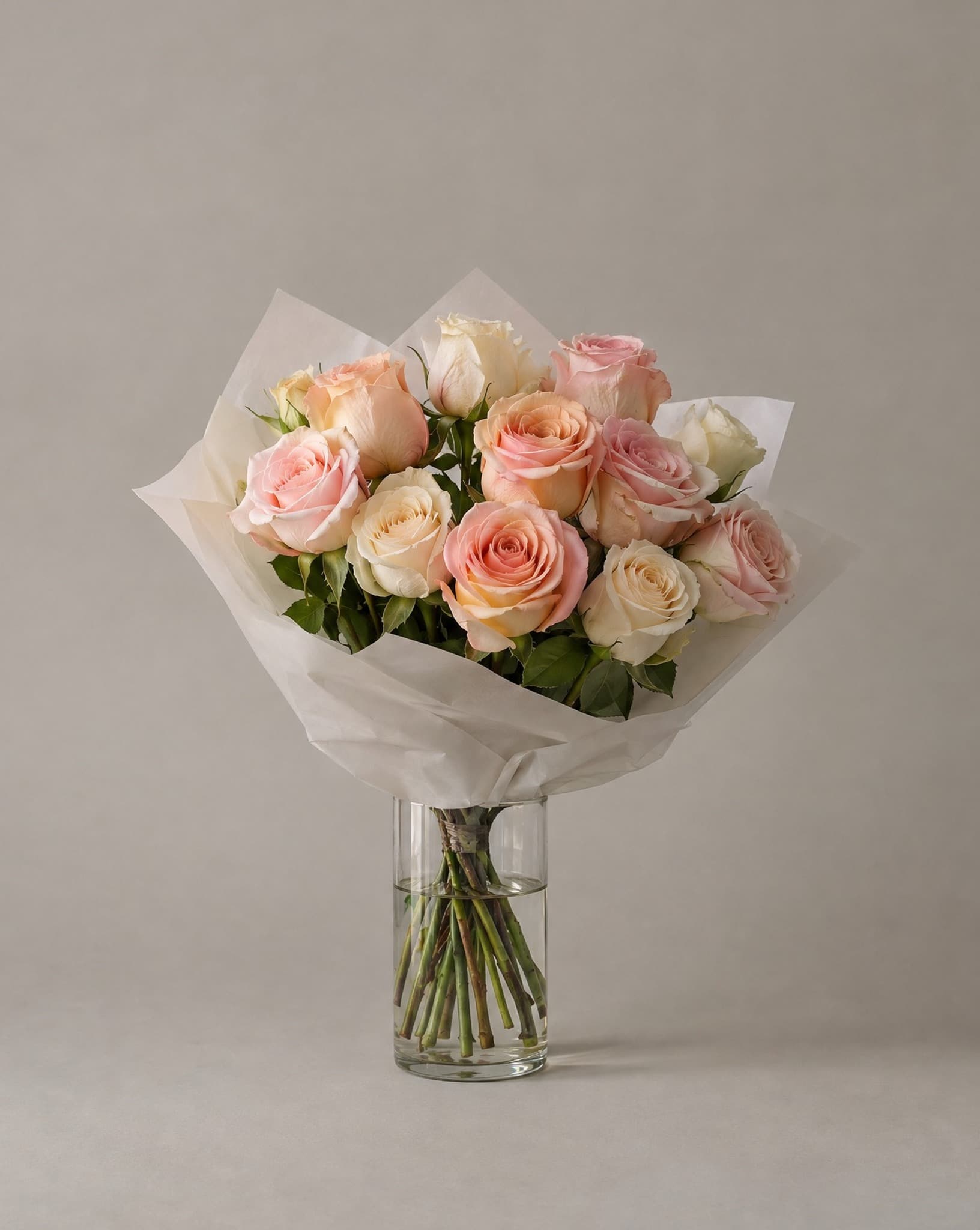 A dozen blush, peach and cream roses hand-wrapped in white tissue, standing in a clear glass vase