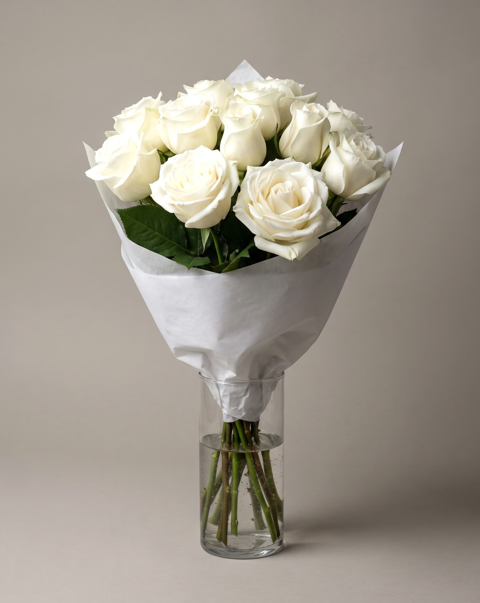 A dozen pure white roses hand-wrapped in white paper, standing in a tall clear glass vase
