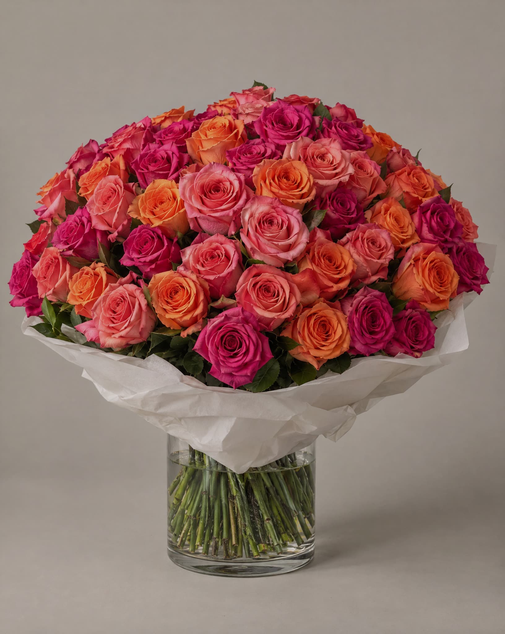 Hot pink, coral and orange roses wrapped in white tissue in a clear glass vase