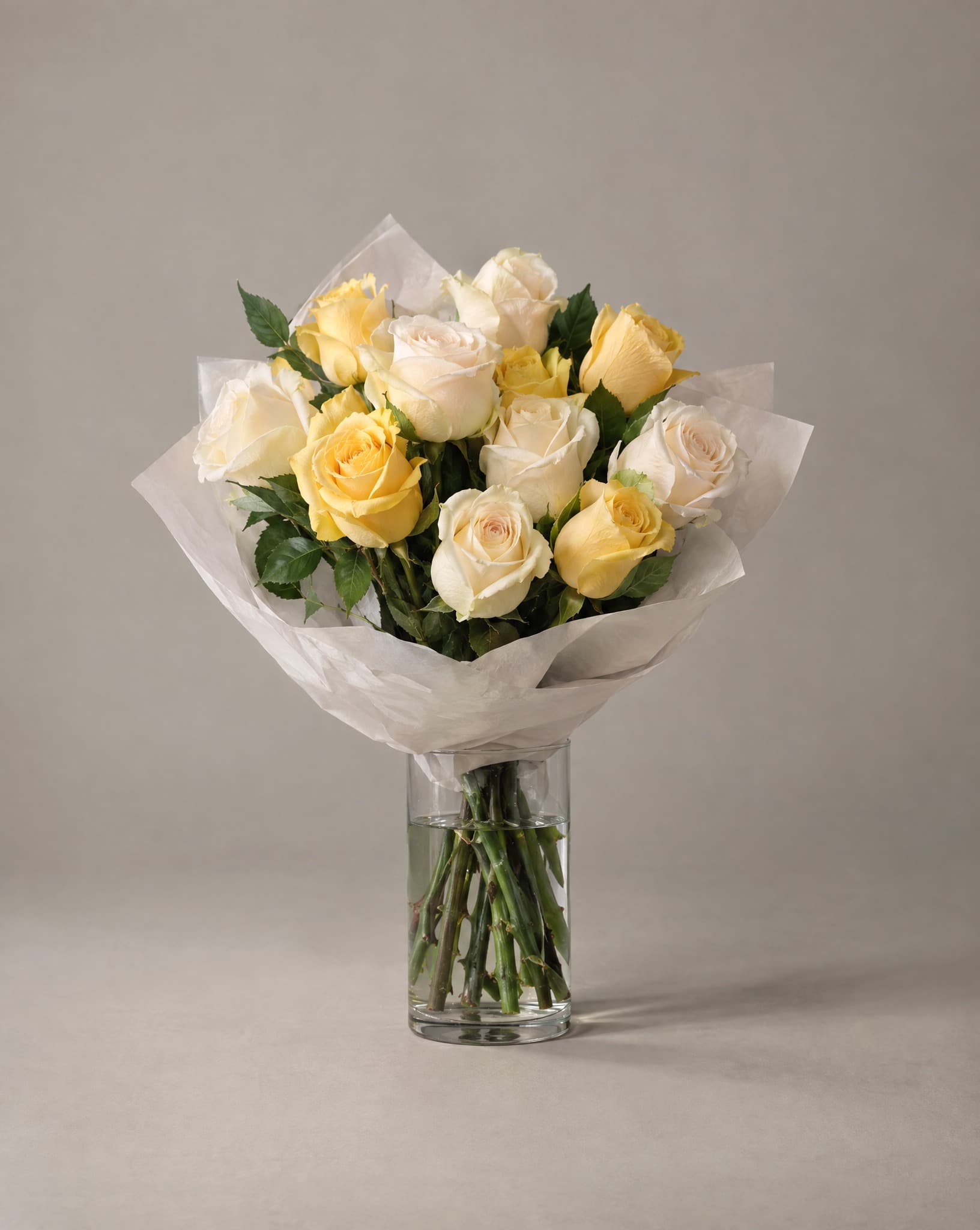 A dozen cream and butter yellow roses hand-wrapped in white tissue, standing in a clear glass vase
