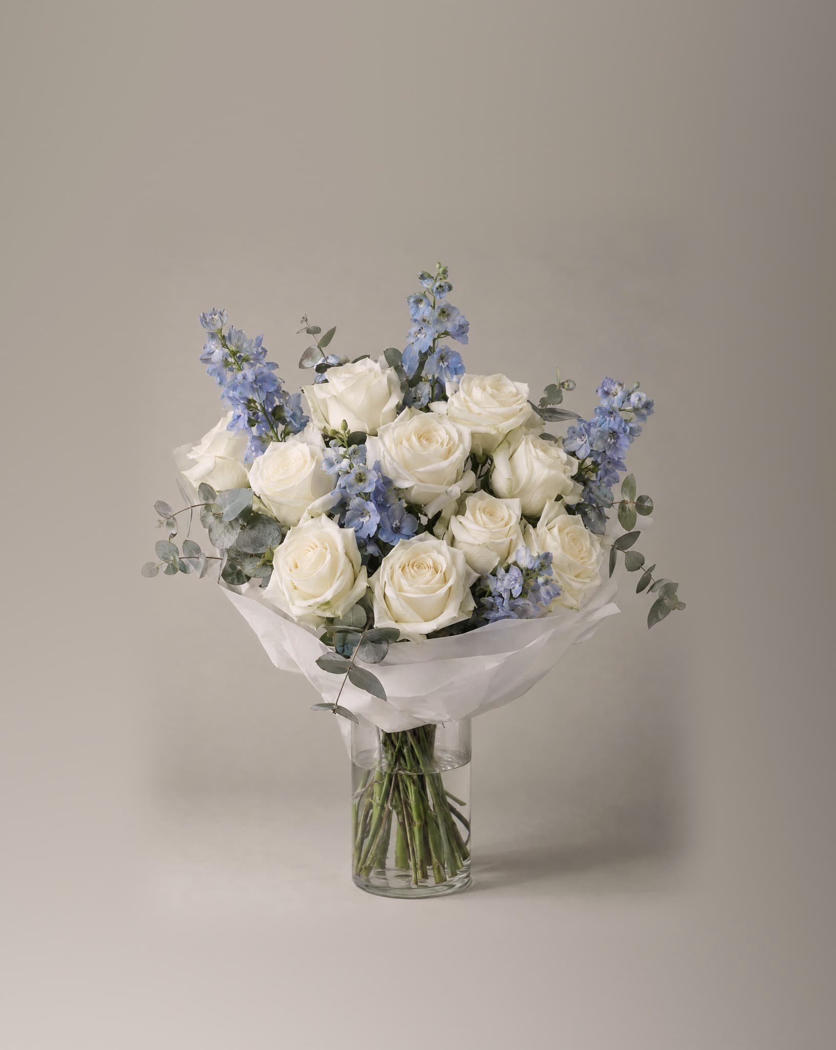 A dozen white roses with pale blue delphinium and eucalyptus, wrapped in white tissue in a clear glass vase