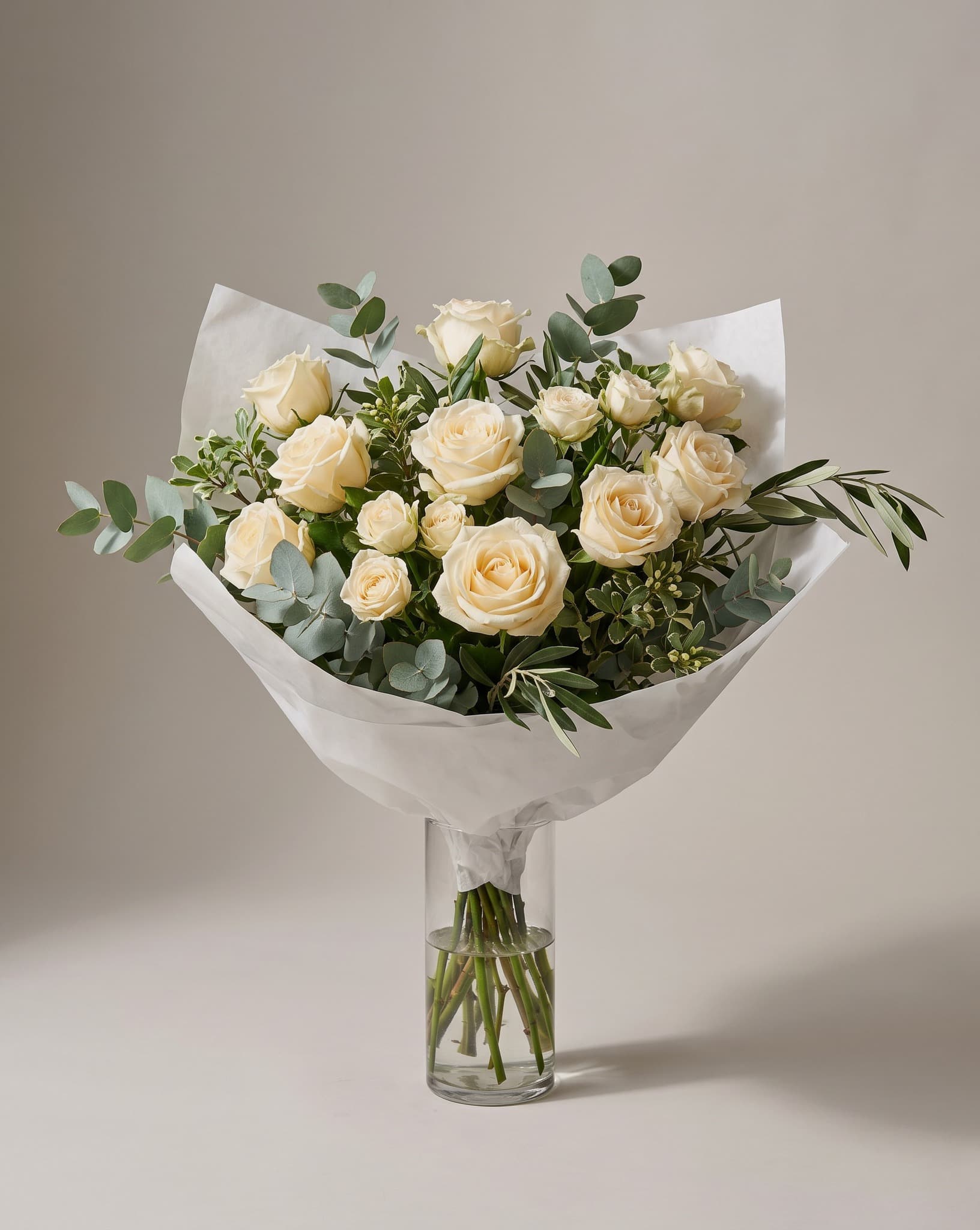 A dozen cream and ivory roses with eucalyptus and olive hand-wrapped in white paper, standing in a tall clear glass vase