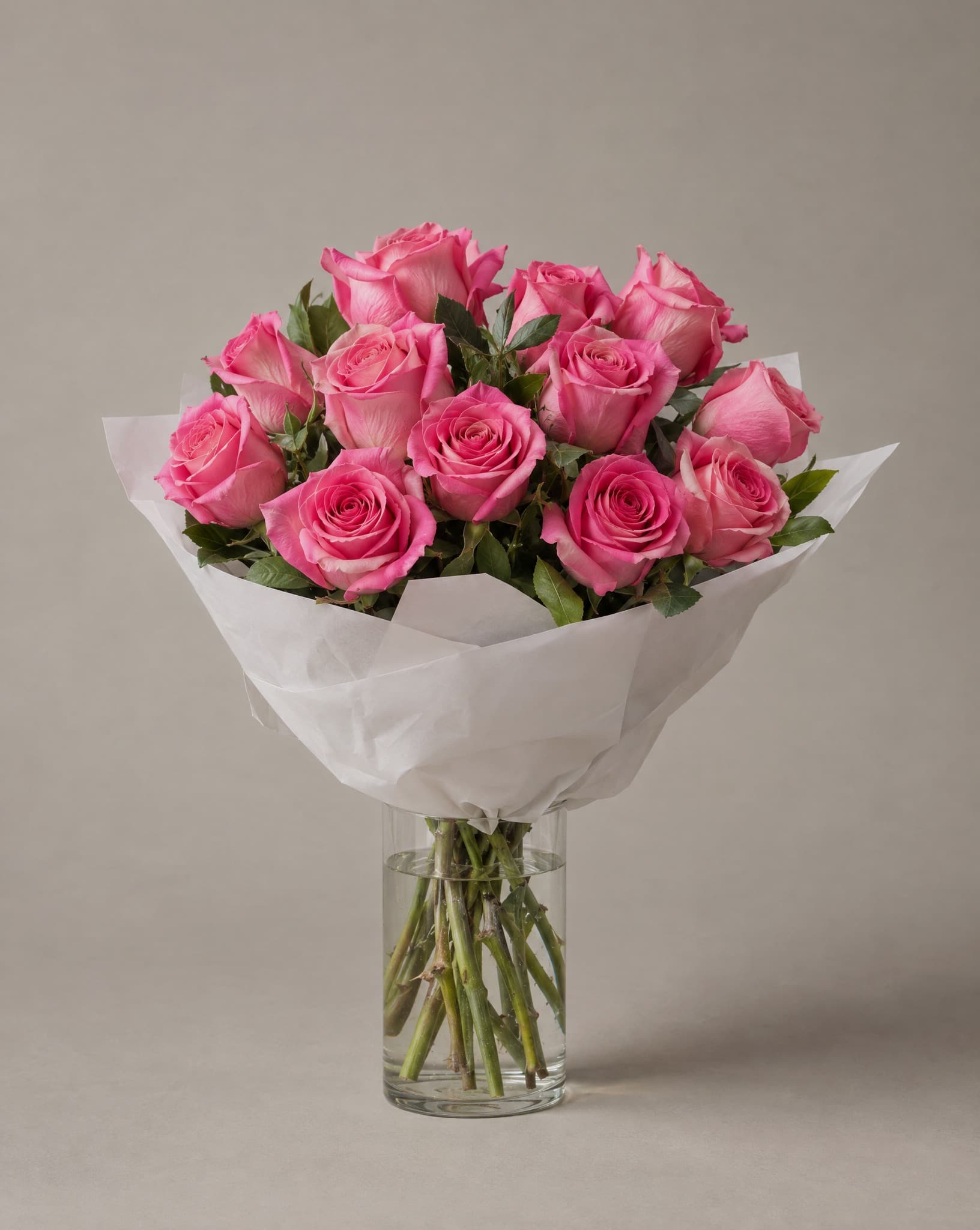A dozen bright pink roses wrapped in white paper in a clear glass vase