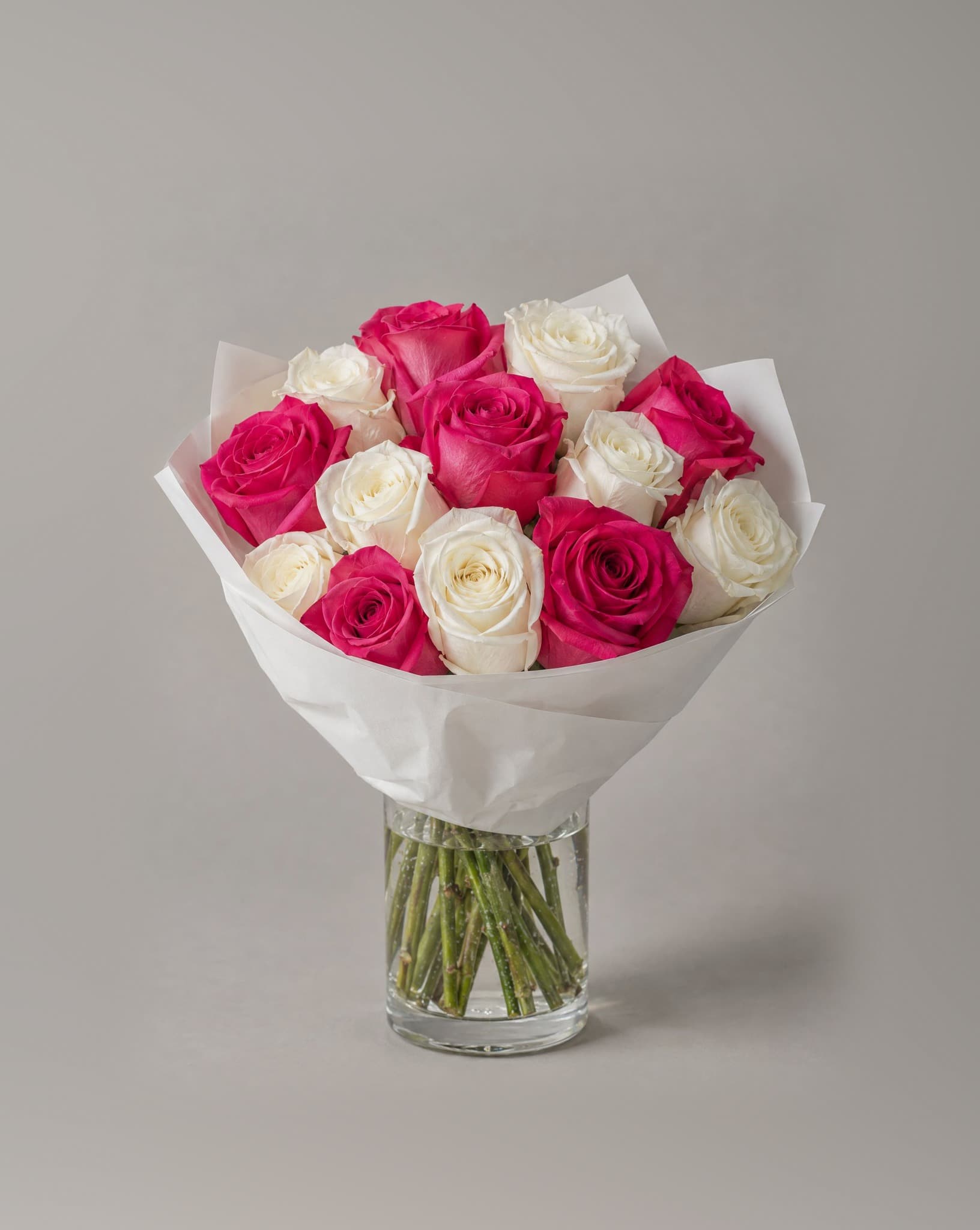 A dozen hot pink and white roses wrapped in white paper in a clear glass vase