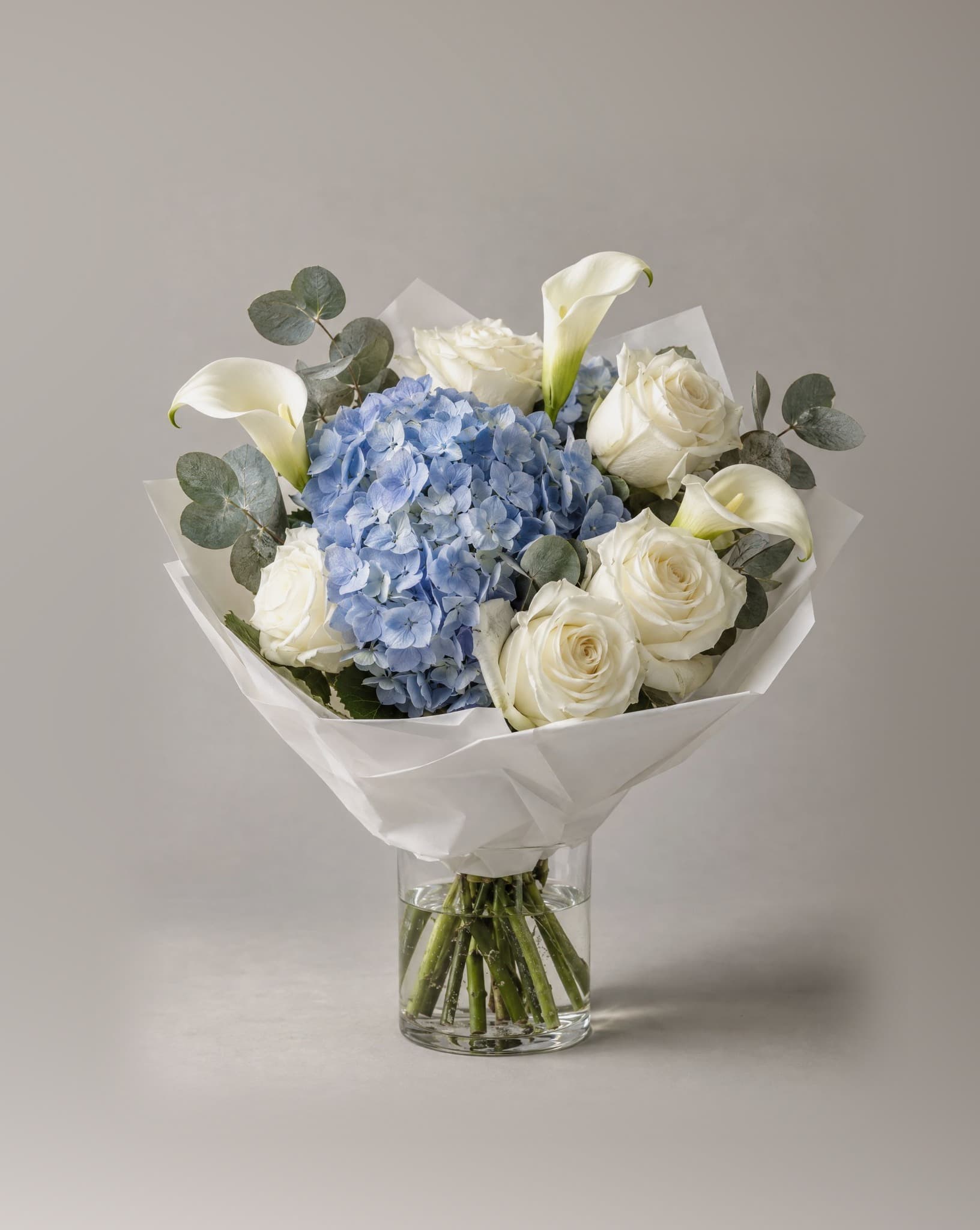 Standard bouquet of blue hydrangeas, white roses and white calla lilies with eucalyptus in a clear glass vase