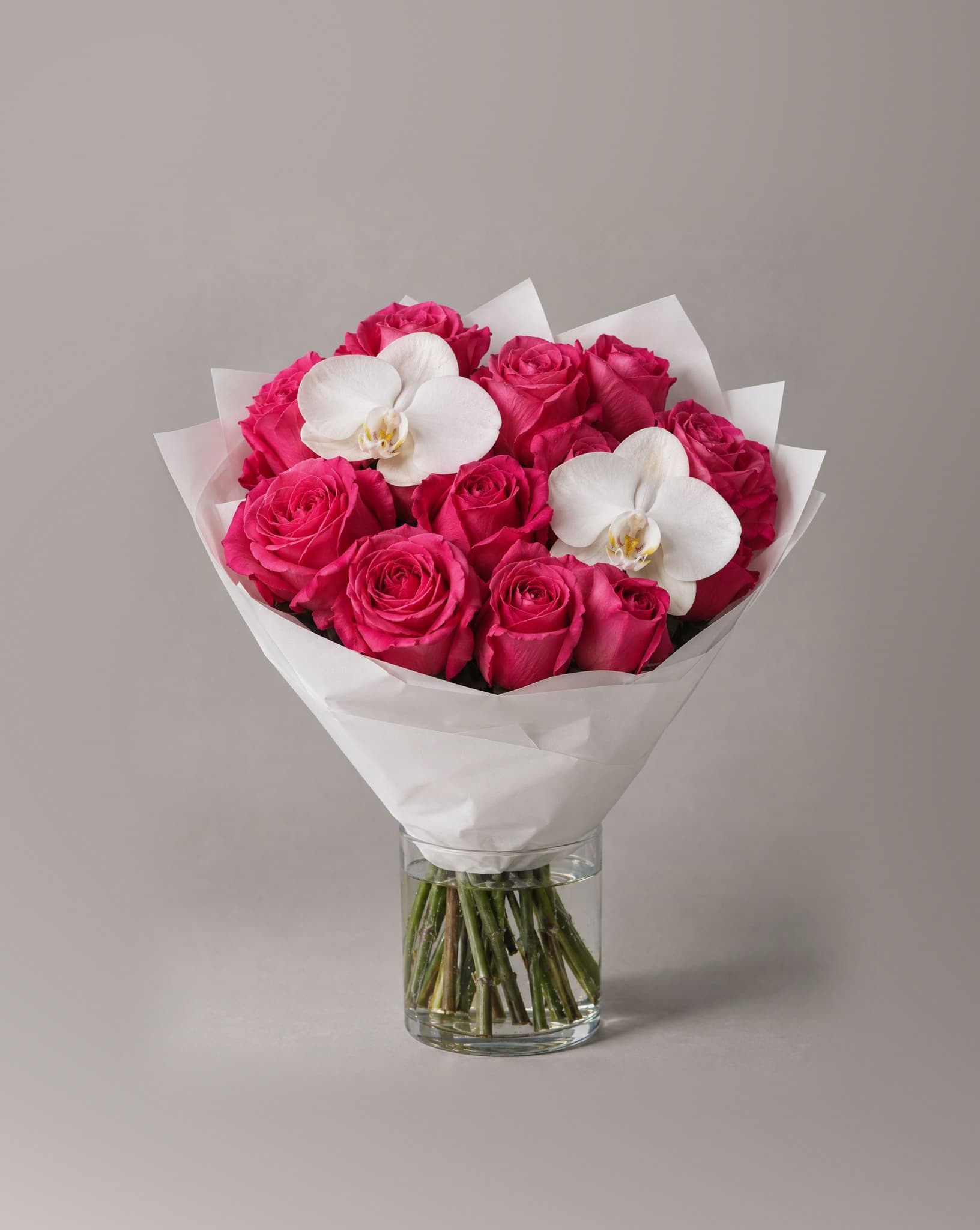 12 hot pink roses with 2 white phalaenopsis orchids wrapped in white paper in a clear glass vase