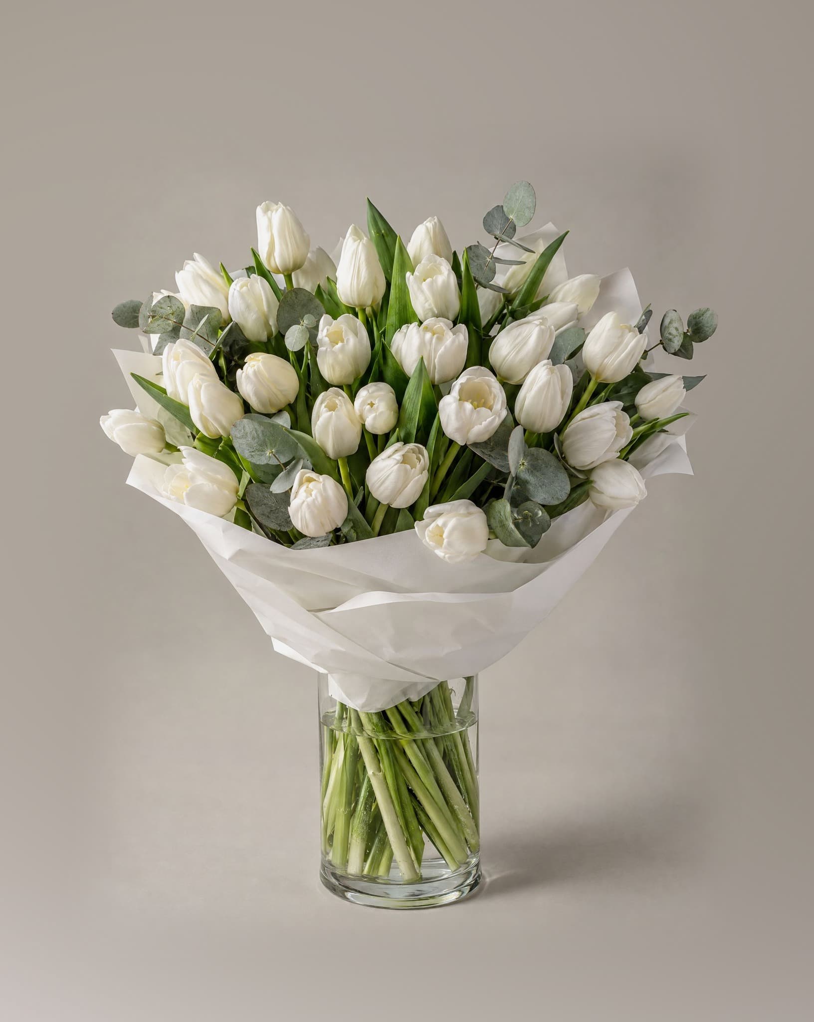 Standard bouquet of white tulips with eucalyptus wrapped in white paper in a clear glass vase