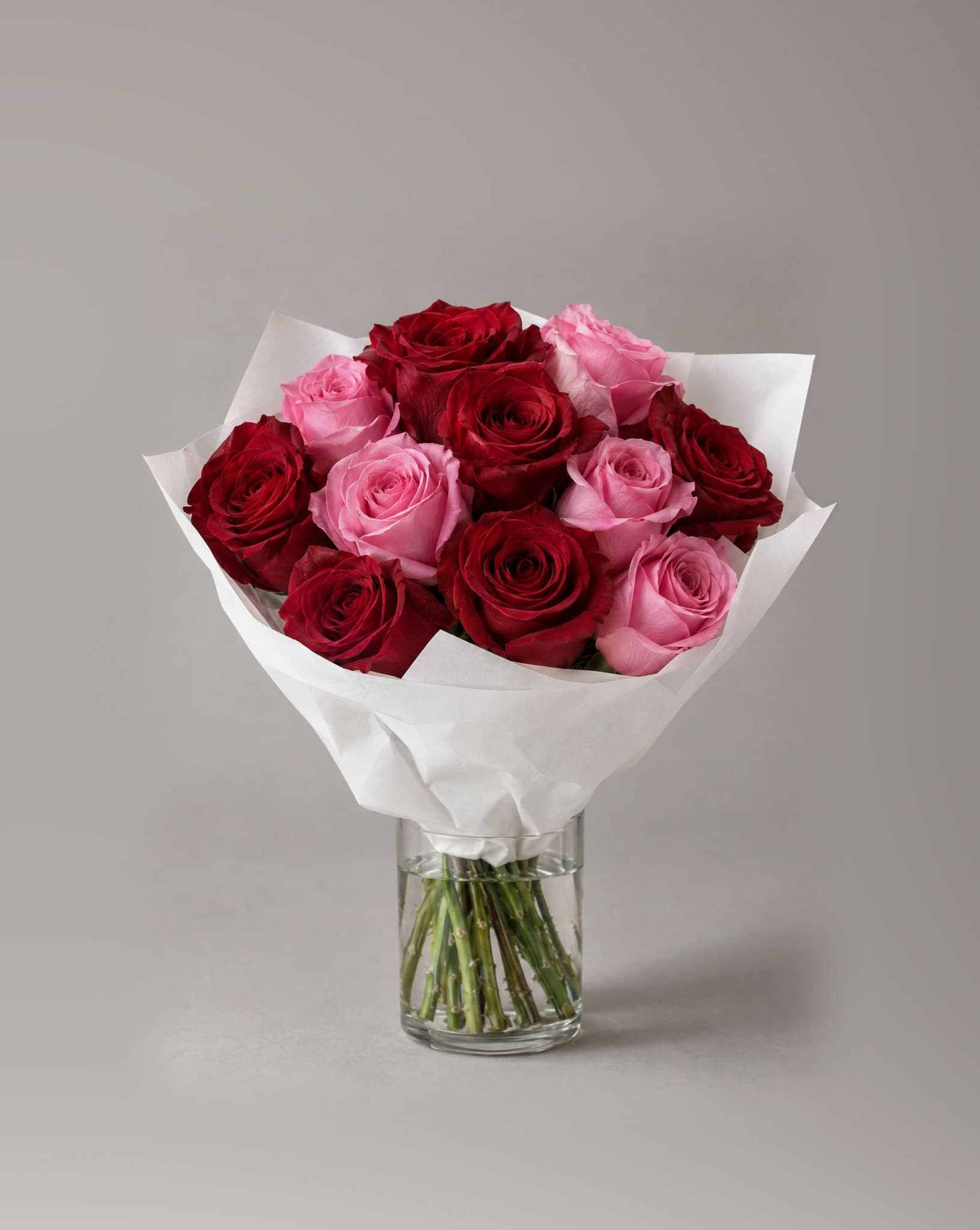 A dozen red and pink roses wrapped in white paper in a clear glass vase
