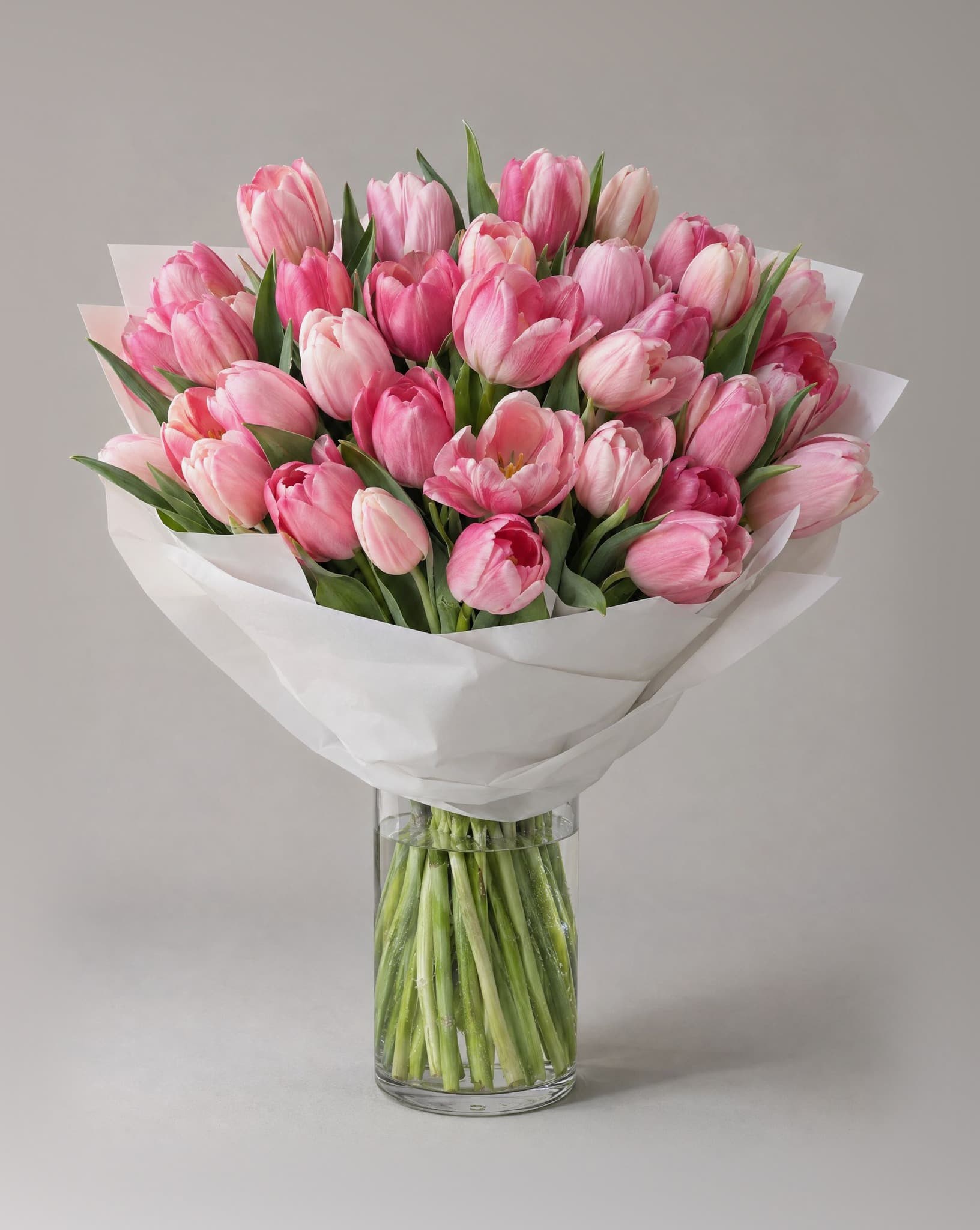 Grand bouquet of pink tulips in shades from blush to rose wrapped in white paper in a clear glass vase